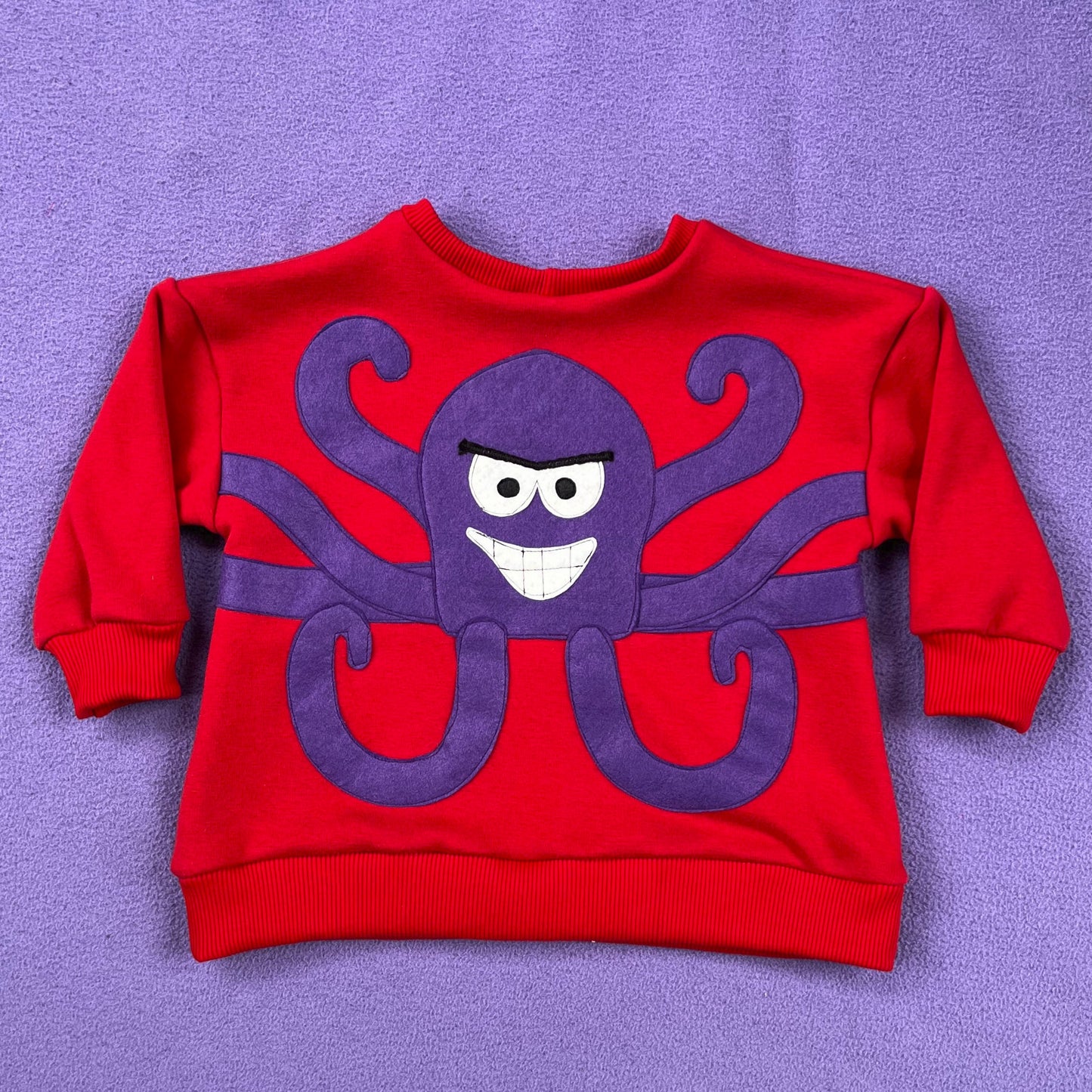 Red, children's sweatshirt with purple Al the Octopus sewn on, on a purple background