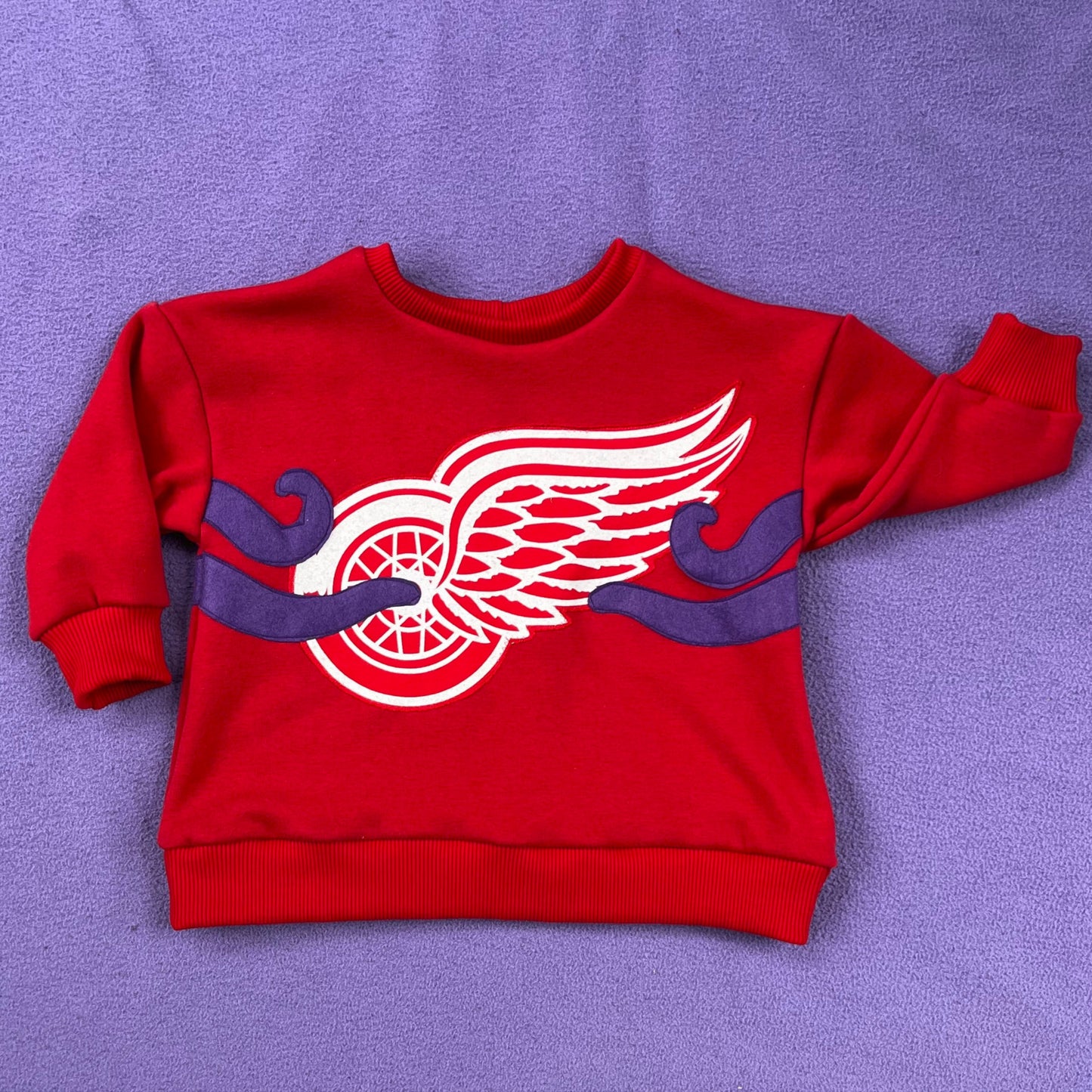 Red, children's sweatshirt with the Red Wings logo and four, purple, octopus tentacles sewn on, on a purple background