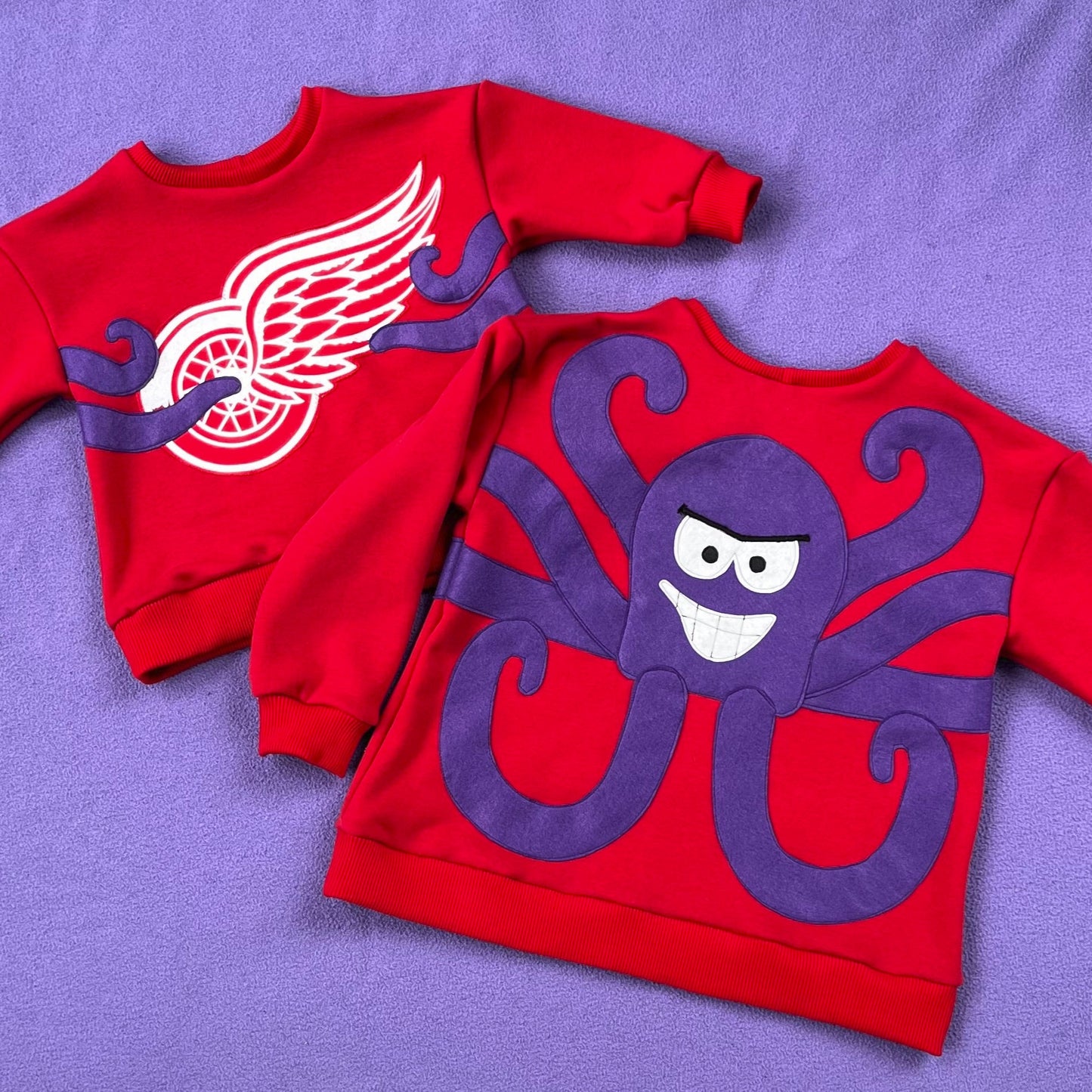 Two red sweatshirts, one with a Red Wings logo and one with Al the Octopus, on a purple background