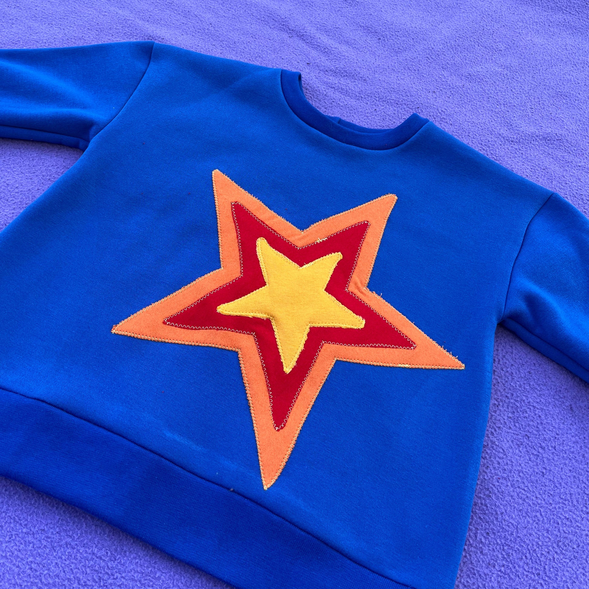 Blue sweatshirt with red, yellow, and orange design on a purple background.