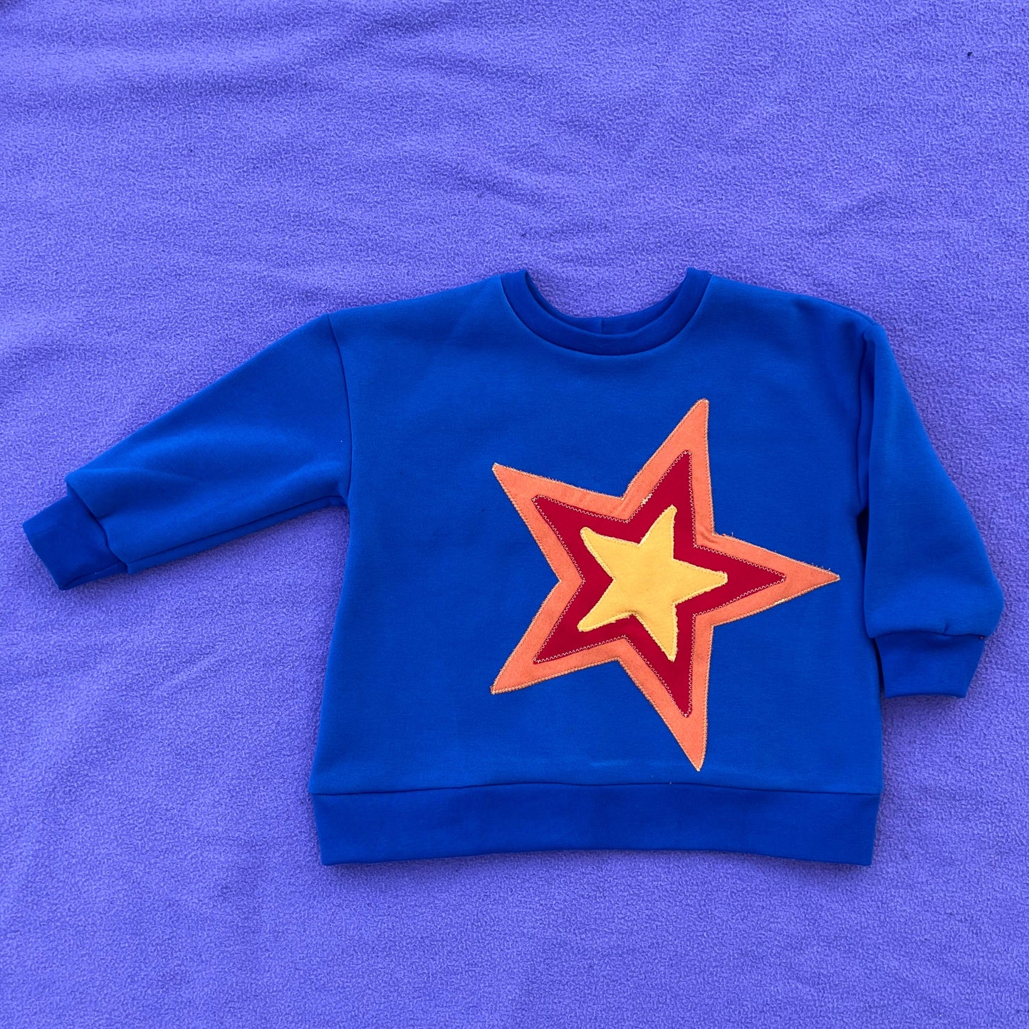Blue sweatshirt with a red, orange, and yellow star design on a purple surface